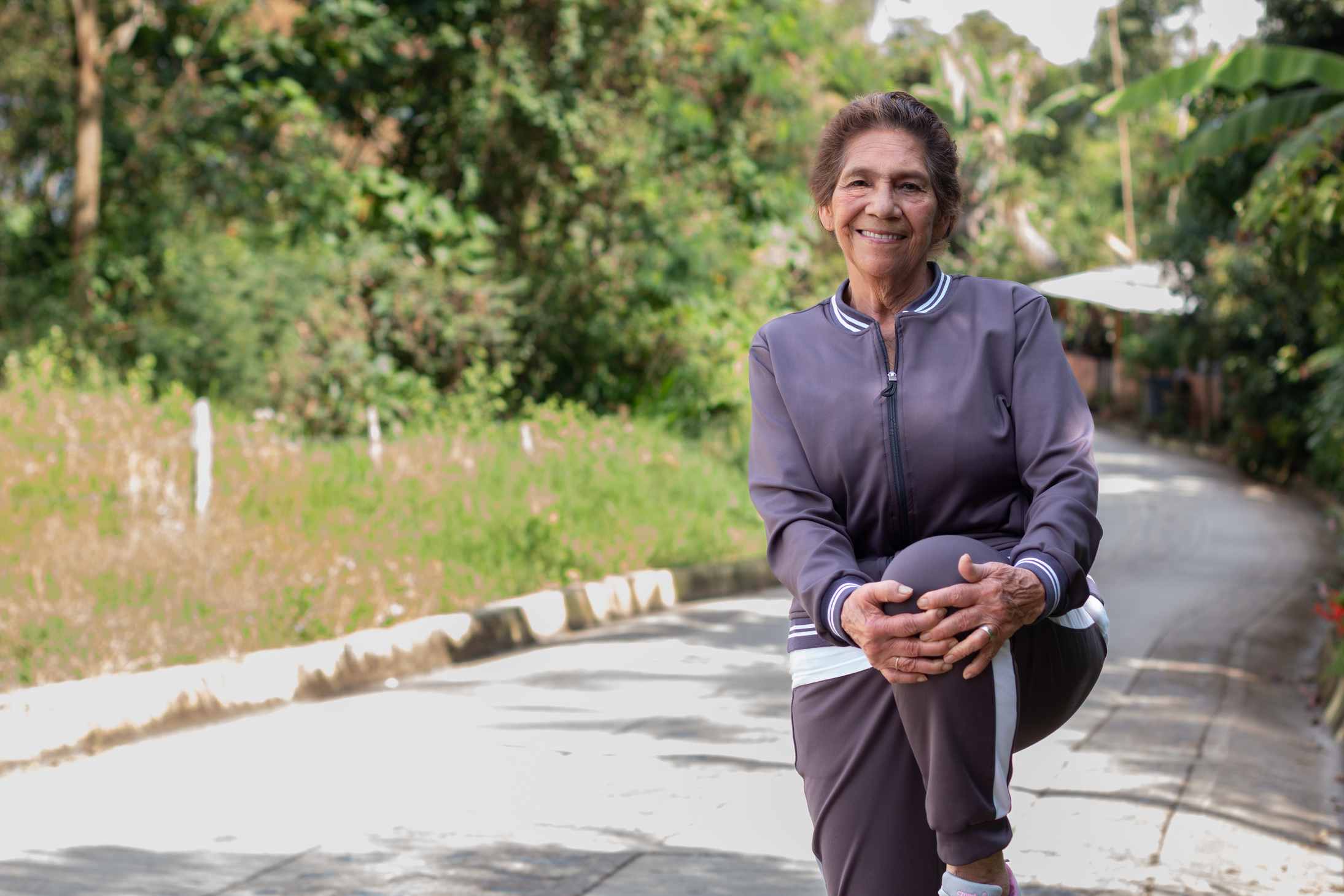 75-year-old woman in sportswear doing knee stretches outdoors, smiling and looking at the camera.